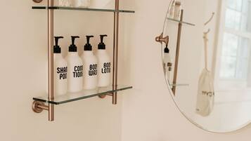 A bathroom with a white vanity, a glass shelf with toiletries, a round mirror, and a brass faucet.