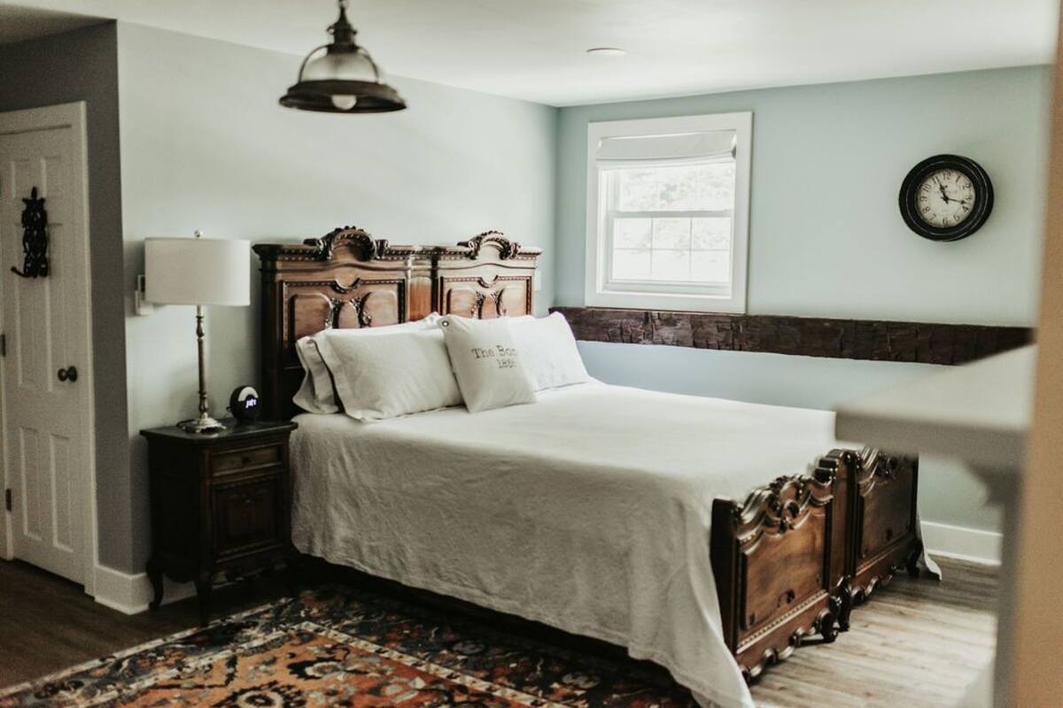 A bedroom with a large bed, a nightstand, a lamp, a clock on the wall, and a window.
