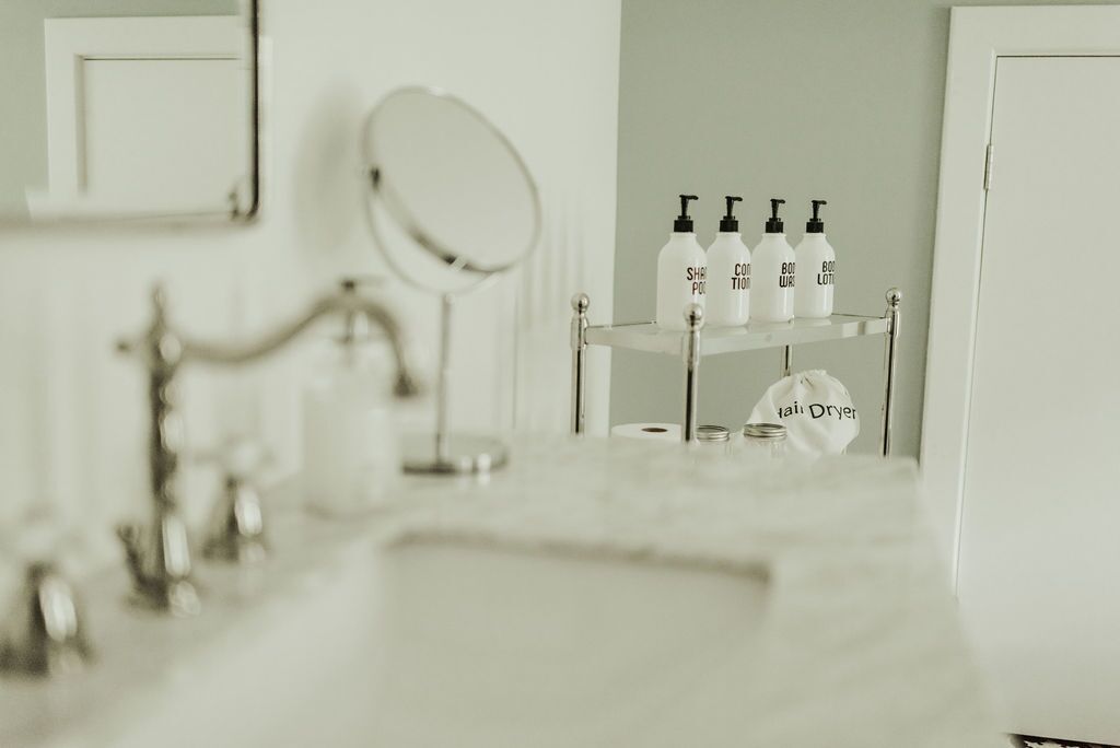 Une salle de bain avec un lavabo blanc, un miroir et une étagère sur laquelle se trouvent des flacons et un sac étiqueté « sèche-cheveux ».