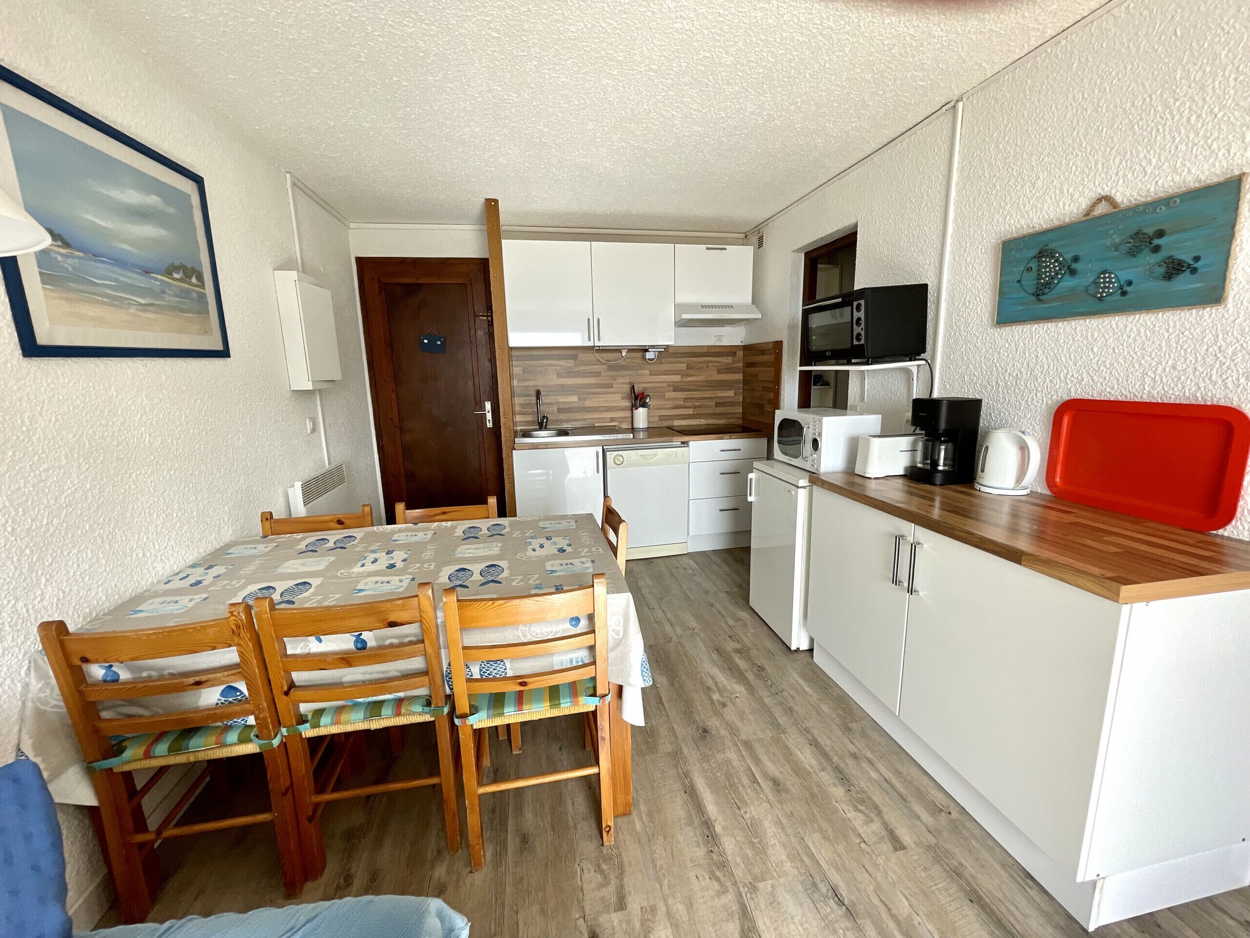 A compact kitchen with white cabinets, a wooden dining table with chairs, a microwave, a kettle, and a red tray.
