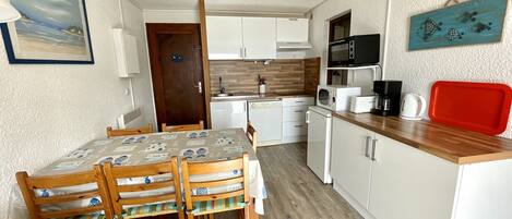 A compact kitchen with white cabinets, a wooden dining table with chairs, a microwave, a kettle, and a red tray.