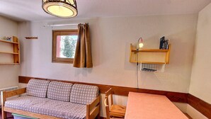 Una habitación con sofá de madera, una mesita con mantel, una ventana con cortinas y un estante en la pared con libros.
