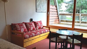 A cozy room with a wooden sofa, a small table, and chairs. There is a window with a view of a wooden deck and trees.