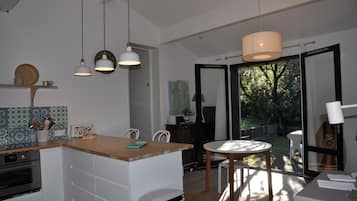 A modern kitchen with white cabinetry, a wooden countertop, and pendant lighting. There is a dining area with a round table and chairs, and a view of a garden through large sliding glass doors.