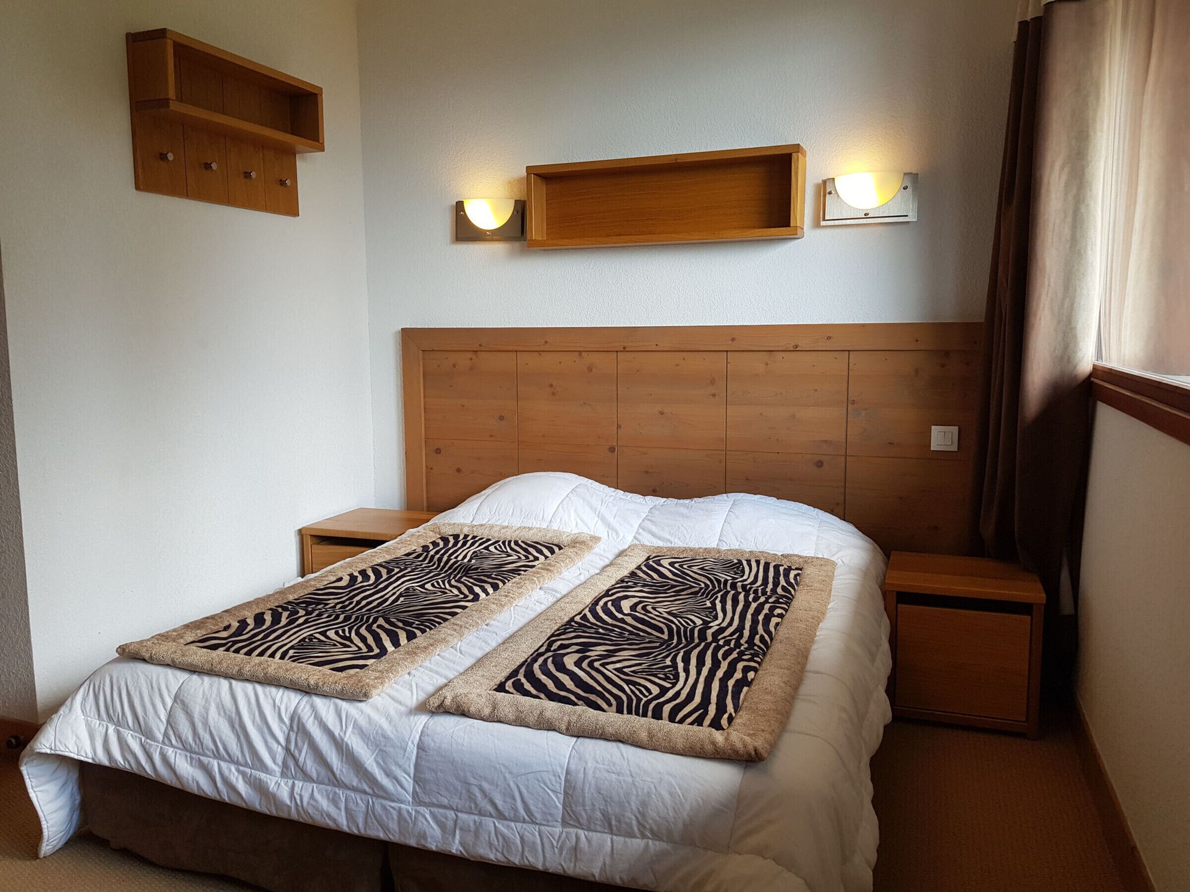 Una camera da letto con testiera in legno, copripiumino bianco, due coperte decorative a motivi zebrati, due comodini e lampade a parete.
