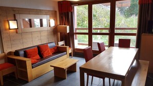 A modern living room with a wooden sofa, a dining table, and chairs. There is a large window with curtains, and a wall-mounted lamp. - 3-room apartment, 8 ppl, Arc 1600, near slopes, pool access & parking (Les Arcs 1600)