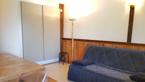Une pièce avec un plancher en bois, un canapé bleu matelassé, une table en bois recouverte d’une nappe en plastique, un petit tabouret et une armoire à portes coulissantes.