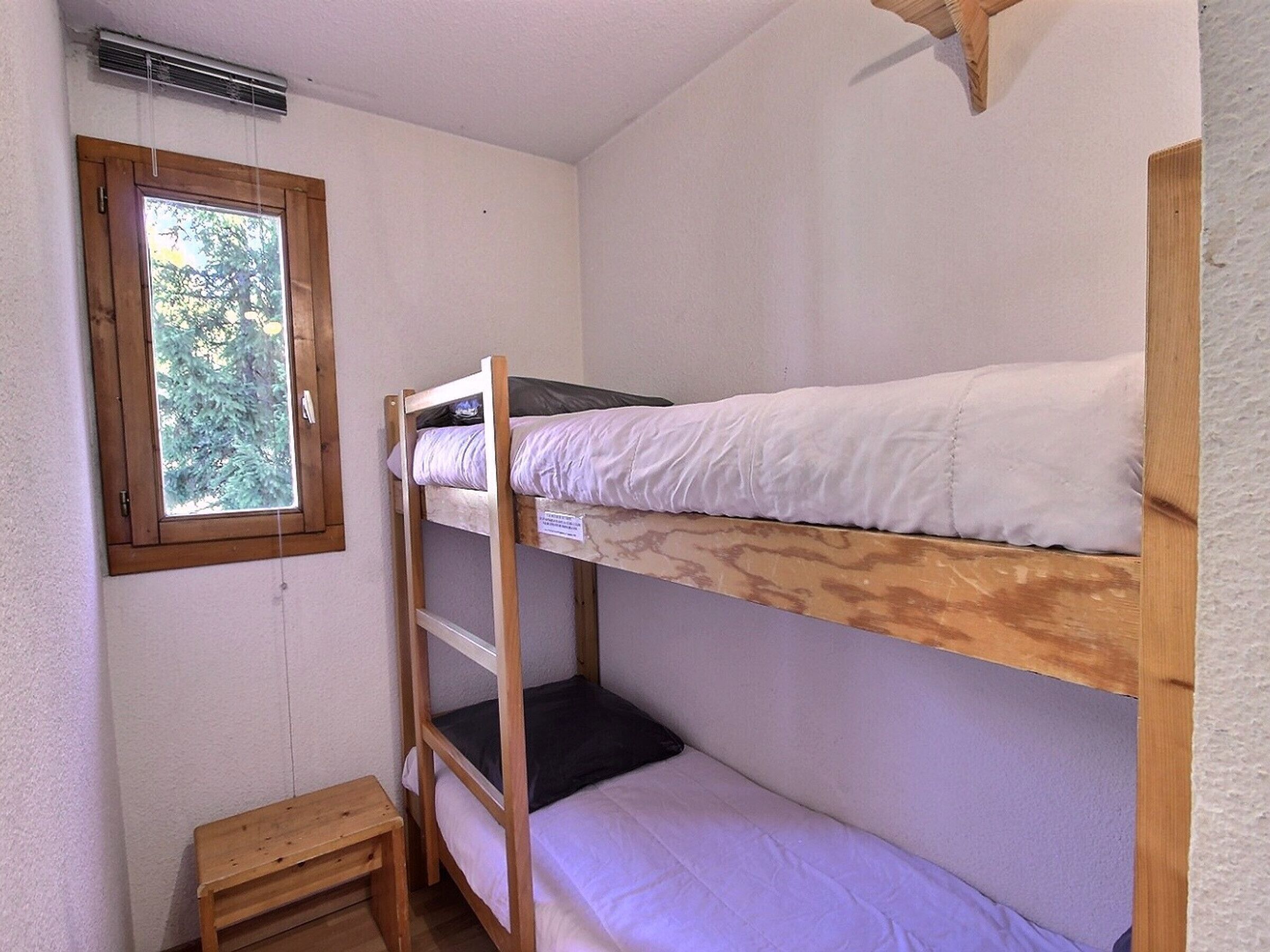 A bunk bed room with a window, a small wooden stool, and a wall-mounted heater.