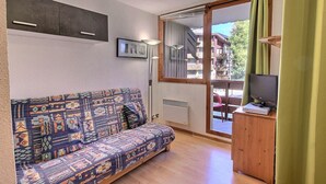 A small living room with a patterned sofa, a television on a wooden stand, and a view of the outside through a sliding glass door. - Cozy studio for 4 in Vallandry, close to slopes (Peisey-Vallandry)
