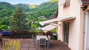A balcony with a table and chairs, a view of a green hillside, and a building with a tiled roof. - Homerez - Apt in Oderen with mountain view (Oderen)