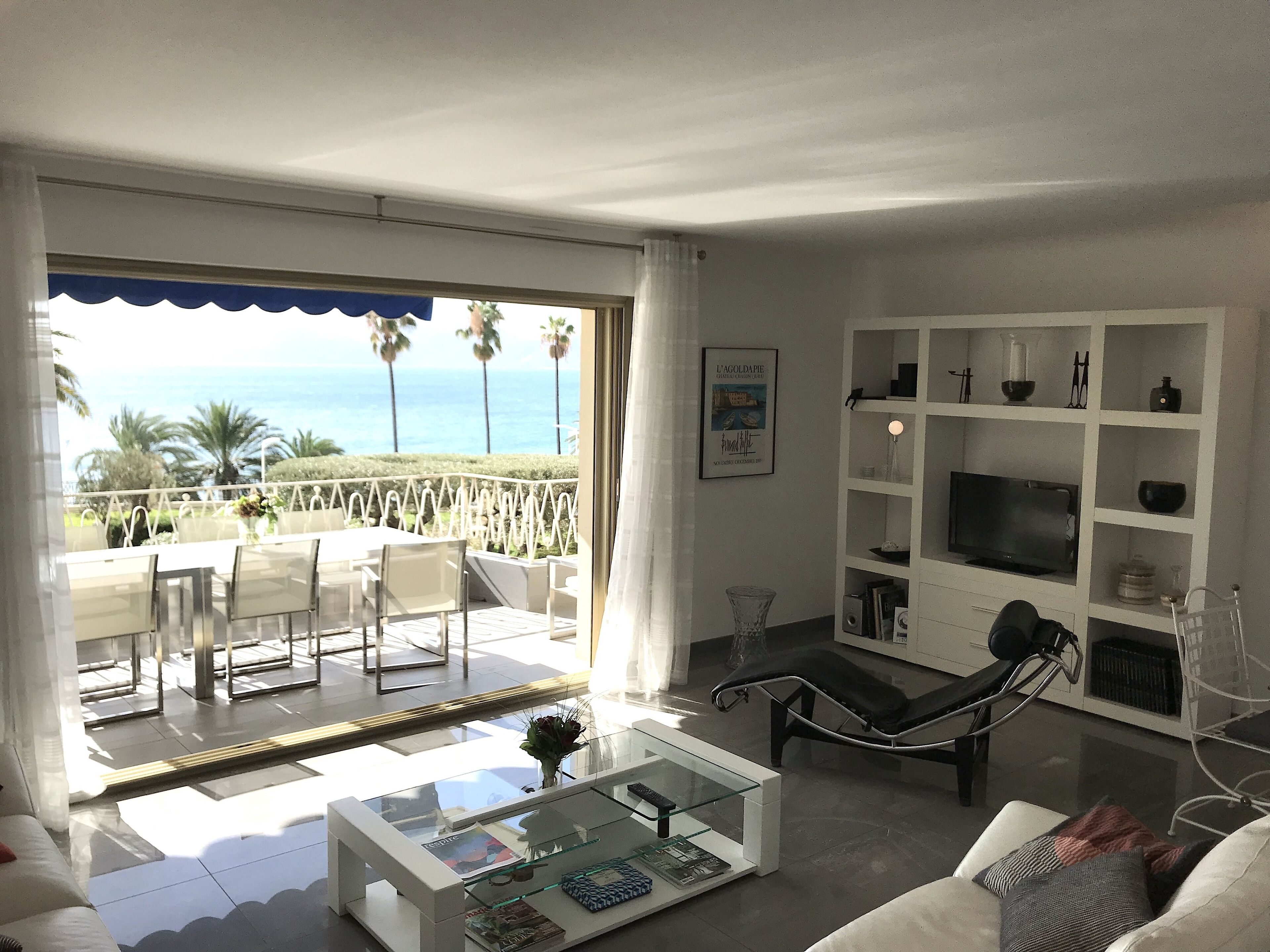 A living room with a glass coffee table, white sofas, a TV, and a bookshelf. A balcony with a view of the ocean and palm trees is visible.