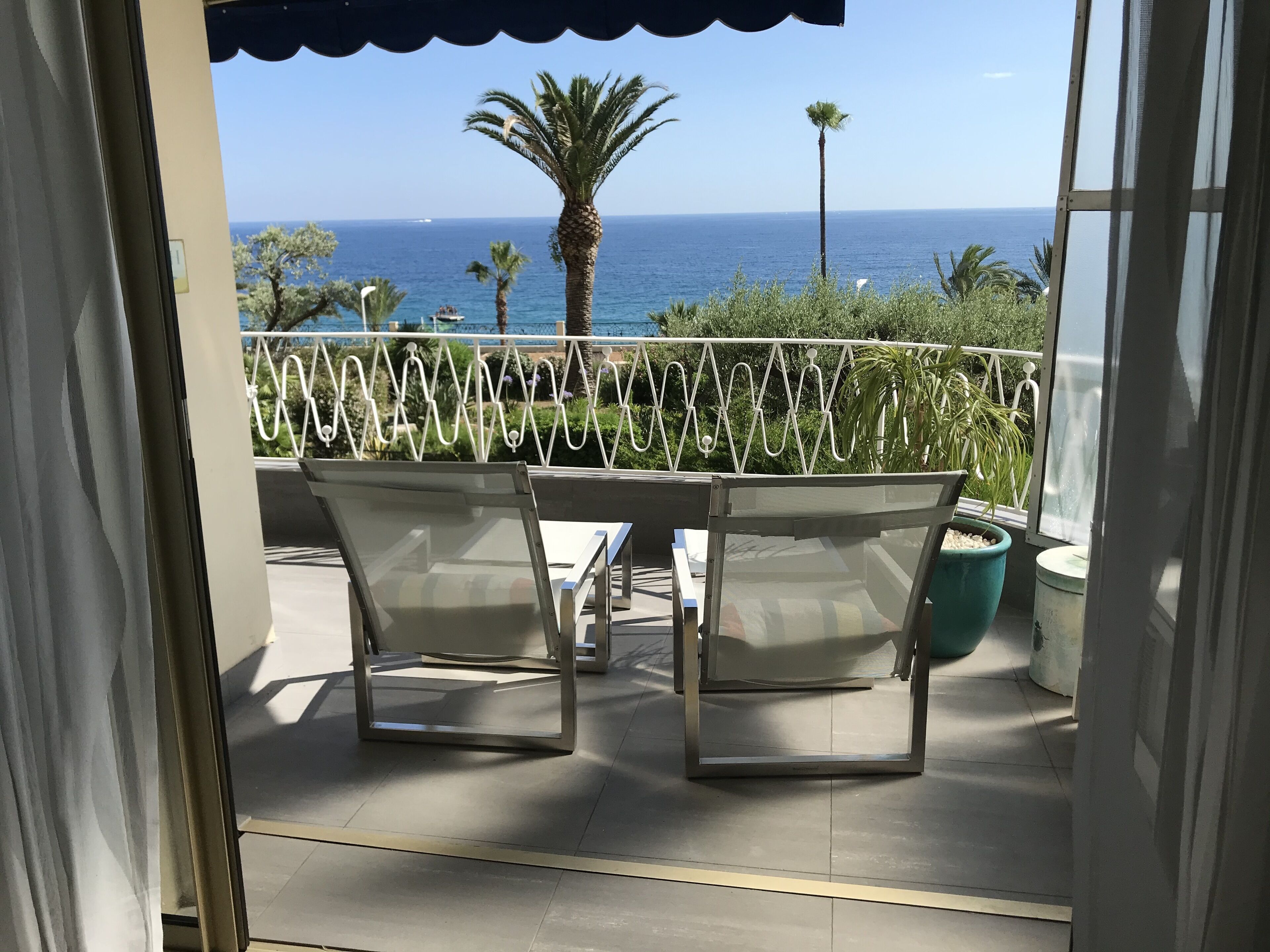 Balcón con dos sillas y una mesa, con vista al mar, palmeras y la costa.