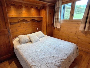 Habitación tipo cabaña de madera con cama, ventana con cortinas y vista a follaje exterior.