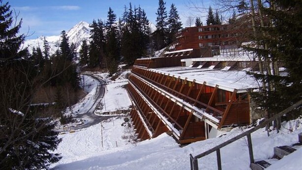 En snötäckt skidlift med en träkonstruktion, omgiven av träd och en bergskedja i bakgrunden.