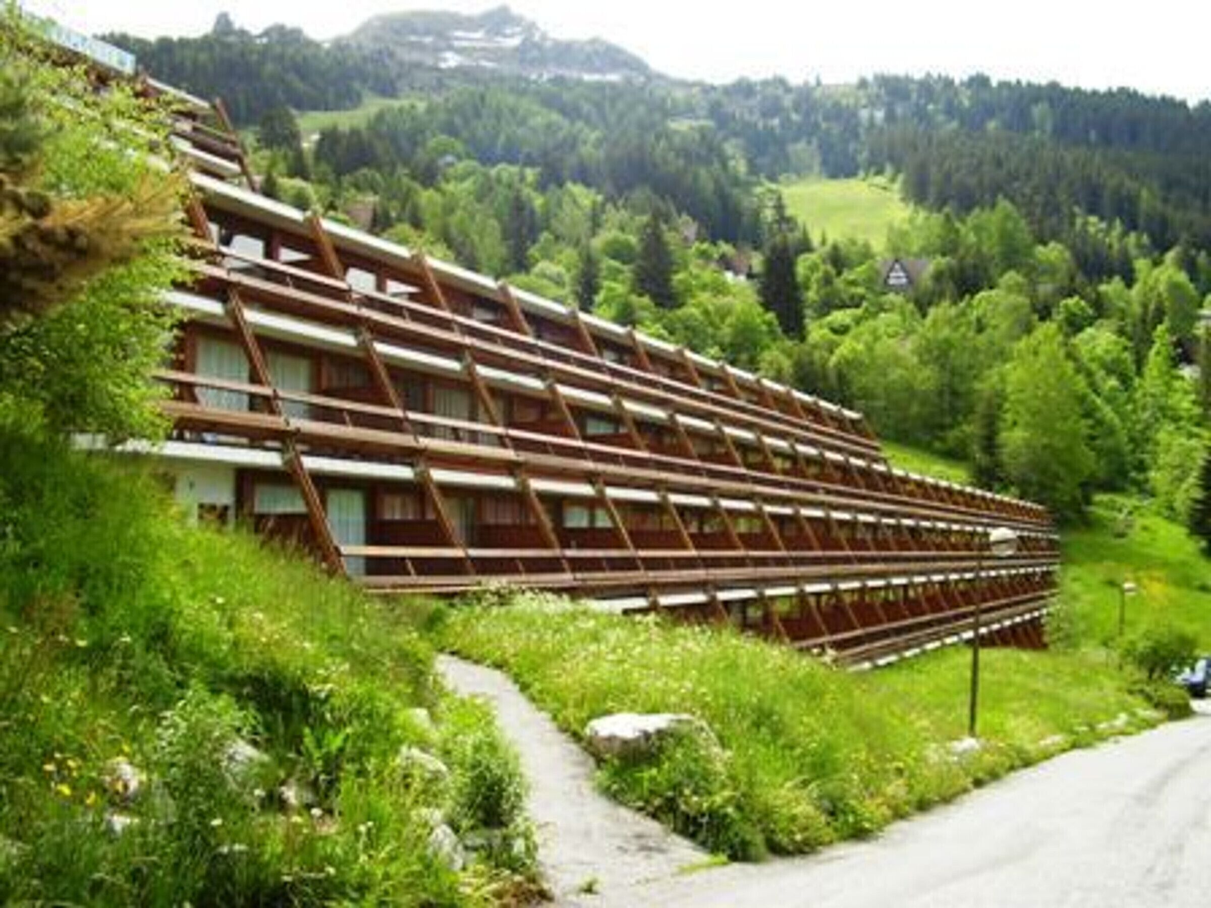 Un edificio grande de varios pisos con techo inclinado, rodeado de vegetación exuberante y un promontorio boscoso.