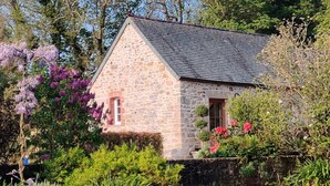 A stone cottage with a sloped roof, surrounded by lush greenery and flowering plants. - Charming house with terrace in Normandy (Grosville)