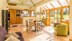 A spacious living area with a dining table, chairs, and a sofa. A kitchen with wooden cabinets and a microwave is visible. Large windows provide natural light and a view of the garden. - Charming house with terrace in Normandy (Grosville)