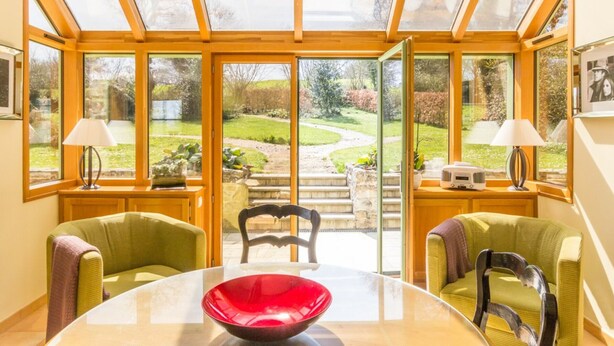 A conservatory with a glass roof, a round table with a red bowl, two lamps, and a view of a garden. - Charming house with terrace in Normandy (Grosville)