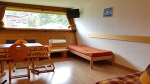 Een kleine, compacte kamer met een houten tafel en stoelen, een enkel bed en een raam met gordijnen.