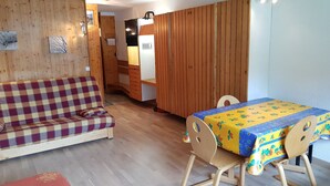 Een knus interieur met een houten vloer, een ruitjesbank, een kleine eettafel met een kleurrijke tafelkleed en een houten kast.