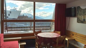 A room with a large window offering a view of mountains and a building, a round table with chairs, a television mounted on the wall, and a red curtain.