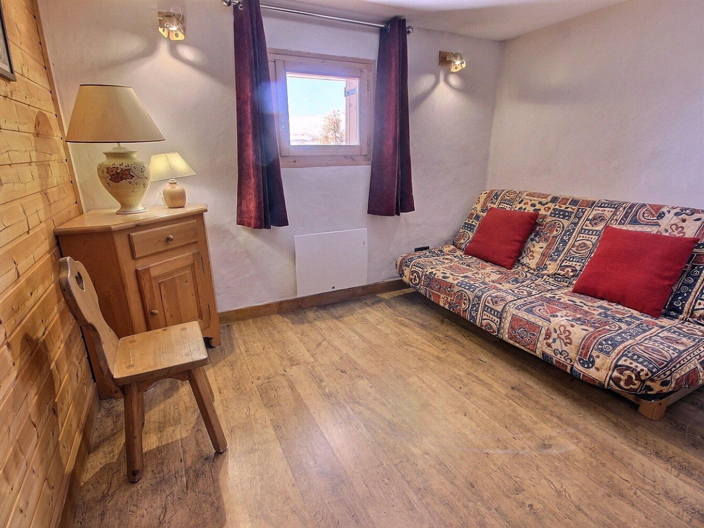 A cozy room with a patterned sofa, a wooden chair, a small table with a lamp, and a window with curtains.