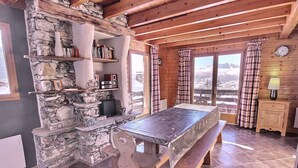 Et hyggeligt sommerhusinteriør med en stenvæggespejs, træbjælker, et spisebord med bænke og udsigt til et sneklædt landskab gennem store vinduer.