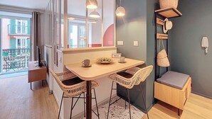 A compact dining area with a wooden table, wicker chairs, and a small bench. A large mirror reflects the room, and a window offers a view of the outside. - N&J - PLACE DU PIN - Port - Close Old Town (Niza)