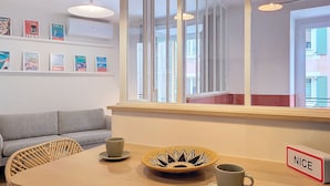 A modern room with a wooden table, wicker chairs, a grey sofa, and a large window with a view of buildings. - N&J - PLACE DU PIN - Port - Close Old Town (Niza)