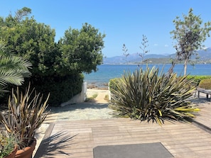 Una terraza de madera con plantas en macetas, un camino que lleva a la playa y vistas al mar y a las montañas.