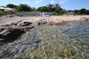 Ein felsiger Strand mit klarem, flachem Wasser, einem Boot und einem Weg zu einem Gebäude.