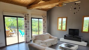 Una sala de estar con un sofá tapizado, un televisor sobre un mueble bajo y un amplio ventanal con vistas a la vegetación.