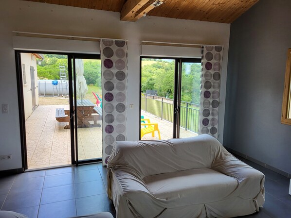 Ein Zimmer mit einer gepolsterten Couch, einer Glasschiebetür, die auf eine Terrasse mit Tisch und Stühlen führt, und Blick ins Grüne.