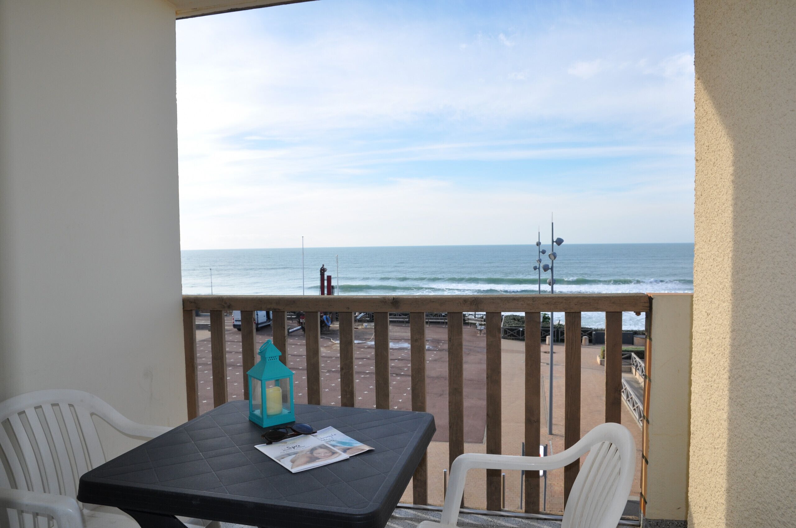 A balcony with a table and chairs, overlooking a beach with a wooden railing and a clear sky.