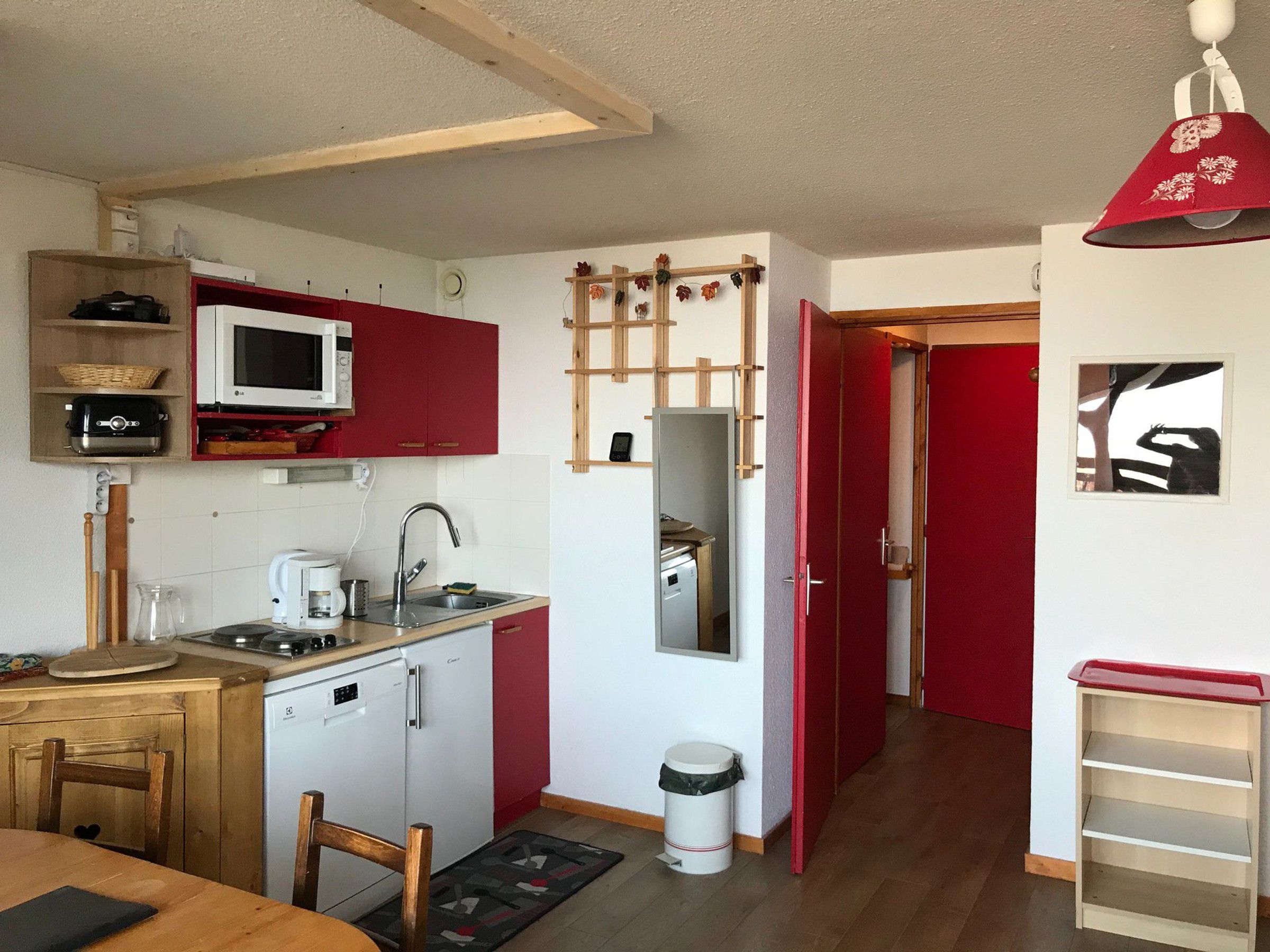 A compact kitchen with a microwave, sink, and small refrigerator. There is a wooden dining table with chairs, a trash can, and a small shelf unit.