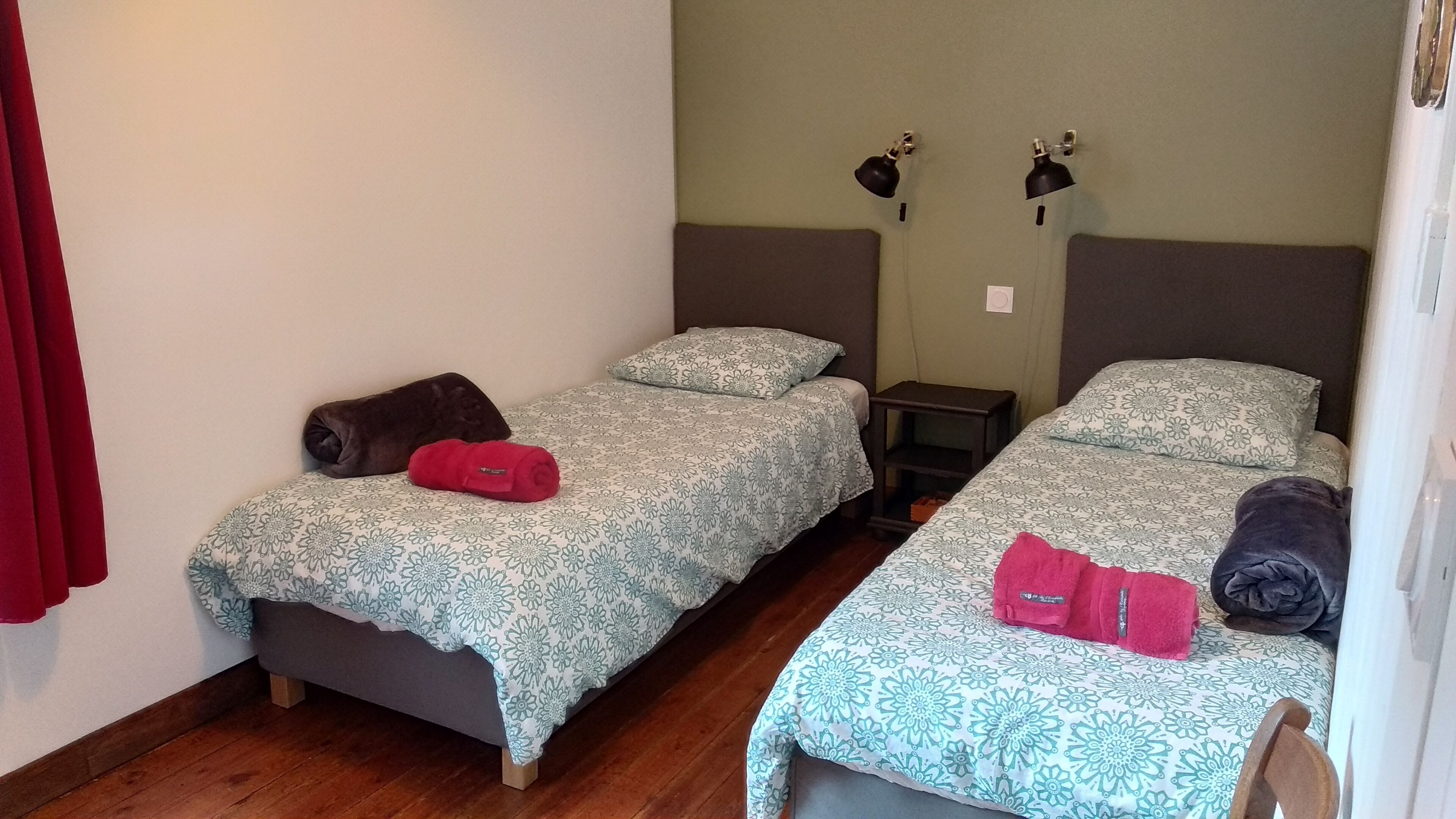 Une chambre avec deux lits simples, chacun agrémenté d’une couvre-lit à motifs et de coussins. Il y a deux tables de chevet, une de chaque côté des lits. La chambre a un plancher en bois et un mur uni avec un interrupteur.