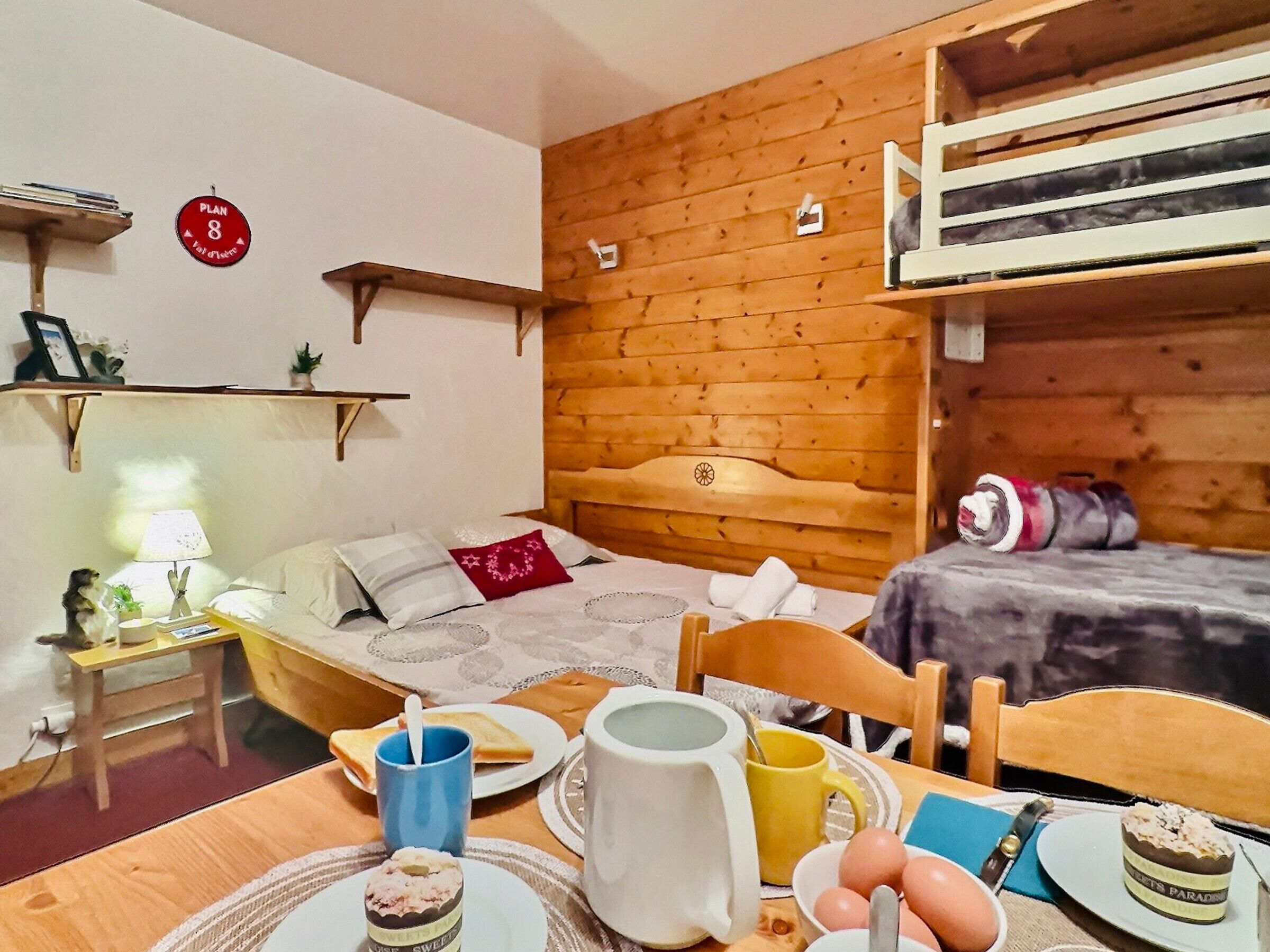 A cozy room with a wooden headboard bed, a small table with breakfast items, a shelf with books, and a 'Plan 8' sign on the wall.