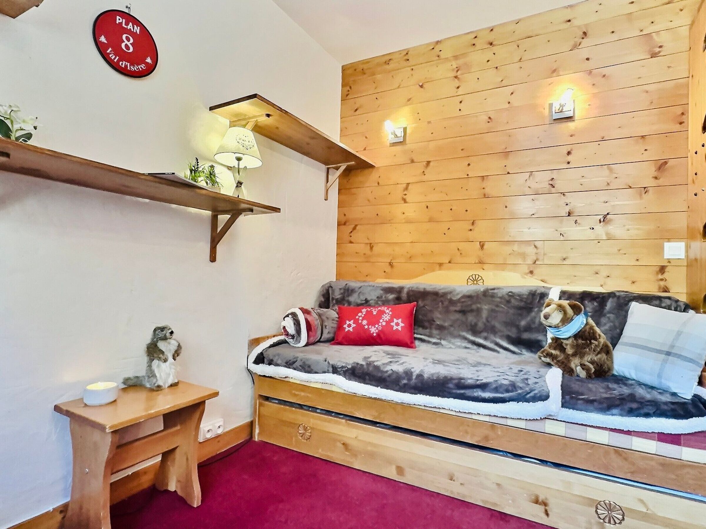 Ein gemütliches Zimmer mit Holzwand, einer Couch mit Kissen, einem kleinen Tisch und einem an der Wand montierten Regal mit einer Lampe.