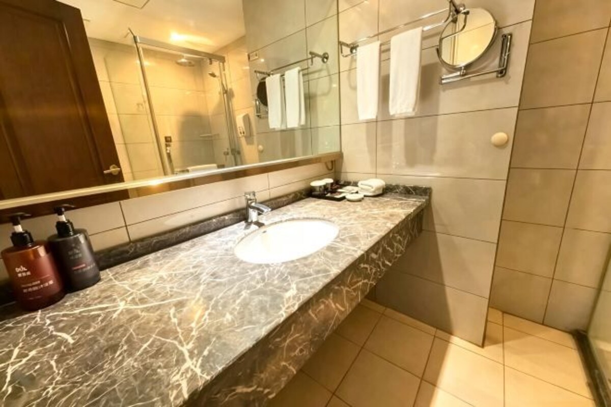 Un baño con lavamanos de mármol, un espejo grande y una ducha visible al fondo.