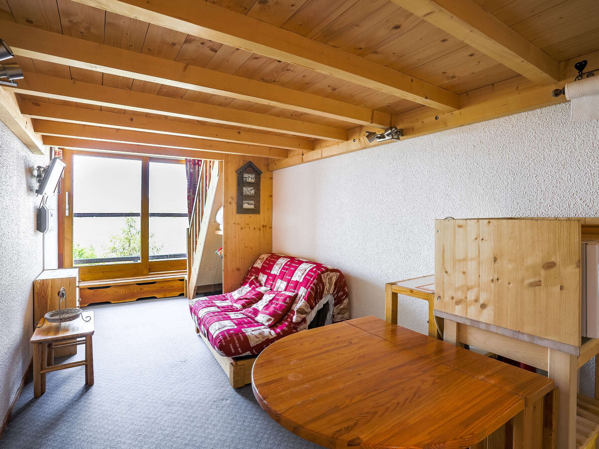 Une pièce chaleureuse avec un plafond en bois, un lit recouvert d’une couette rouge et blanche, une petite table en bois et une fenêtre avec des rideaux.