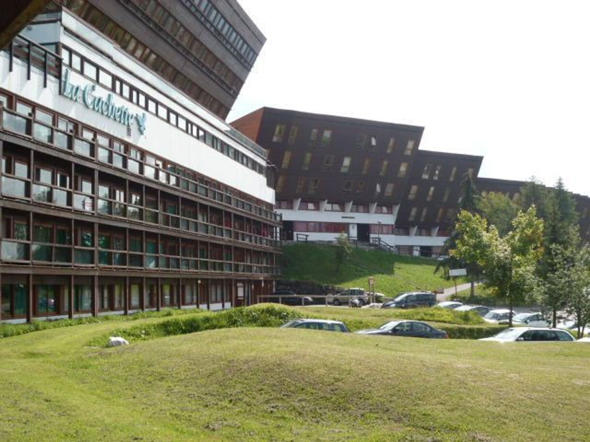 Um edifício moderno com vários andares e telhado plano, cercado por uma área gramada com carros estacionados.
