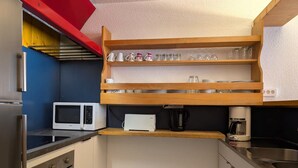 Et kompakt køkken med mikrobølgeovn, kaffemaskine og et hyldesæt med glas.