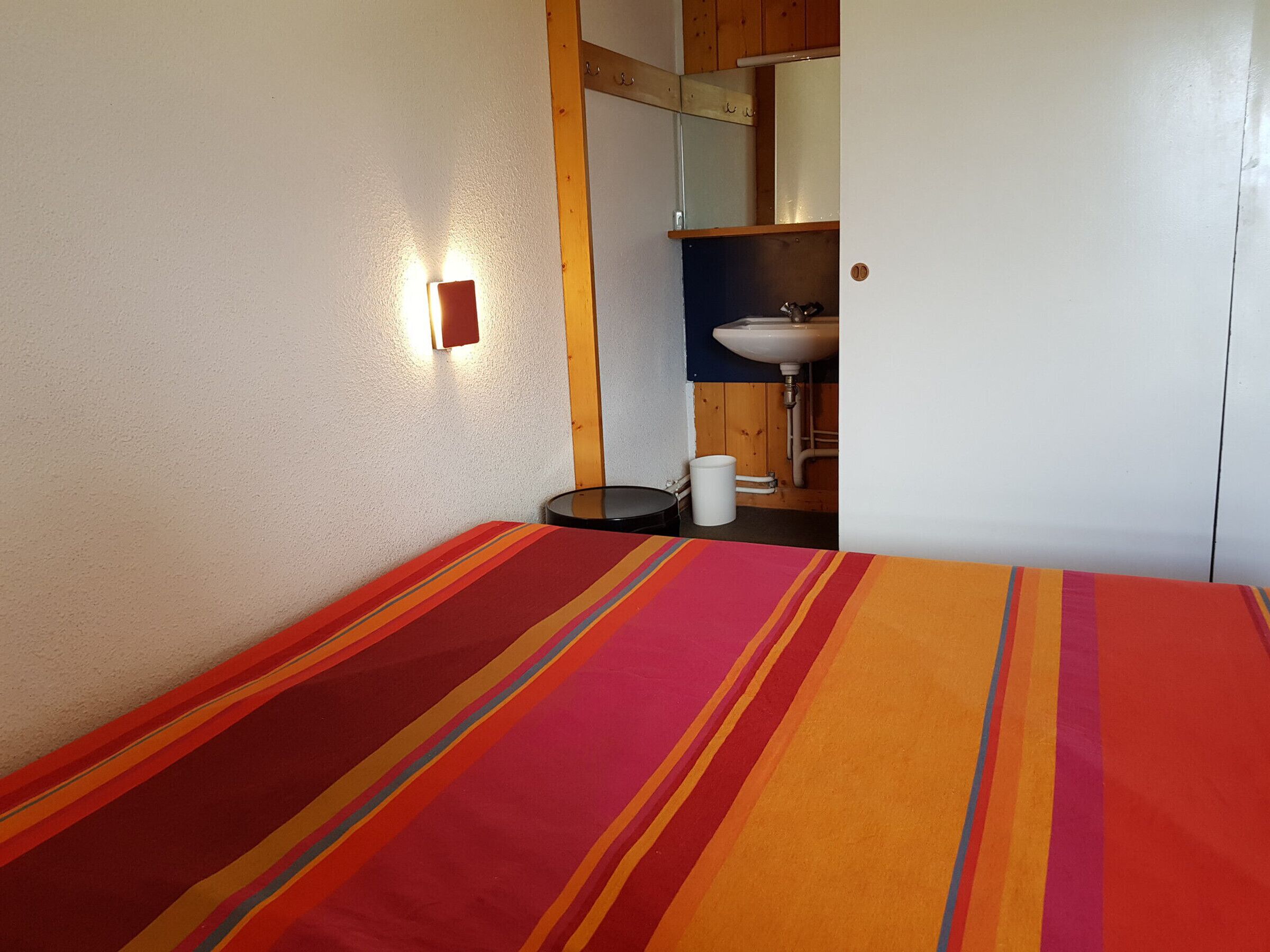 A room with a bed, a small round table, a wall-mounted light, and a bathroom with a sink and mirror.