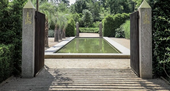 A rectangular pool with a paved walkway leading to it, surrounded by greenery and stone pillars with inscriptions.