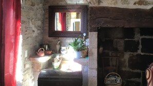 A rustic room with a stone wall, a wooden beam ceiling, a fireplace, a mirror, and a shelf with various items.