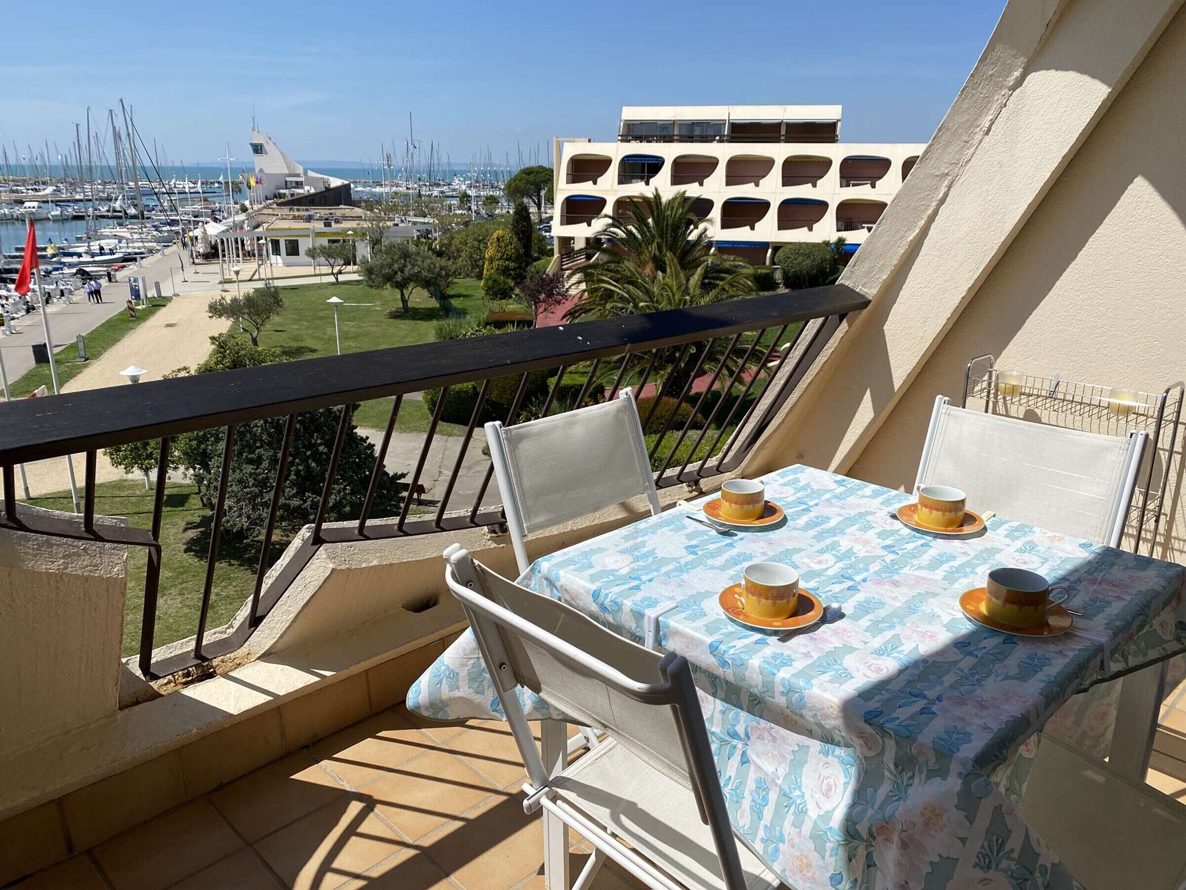 A balcony with a table set for two, overlooking a marina with boats and a building with a unique architectural design.