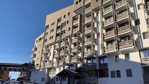 Un hotel a più piani con balconi, una seggiovia sullo sfondo e auto parcheggiate.