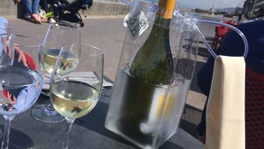 Uma mesa com uma garrafa de vinho branco num recipiente transparente, copos de vinho e vista da praia com pessoas ao fundo.