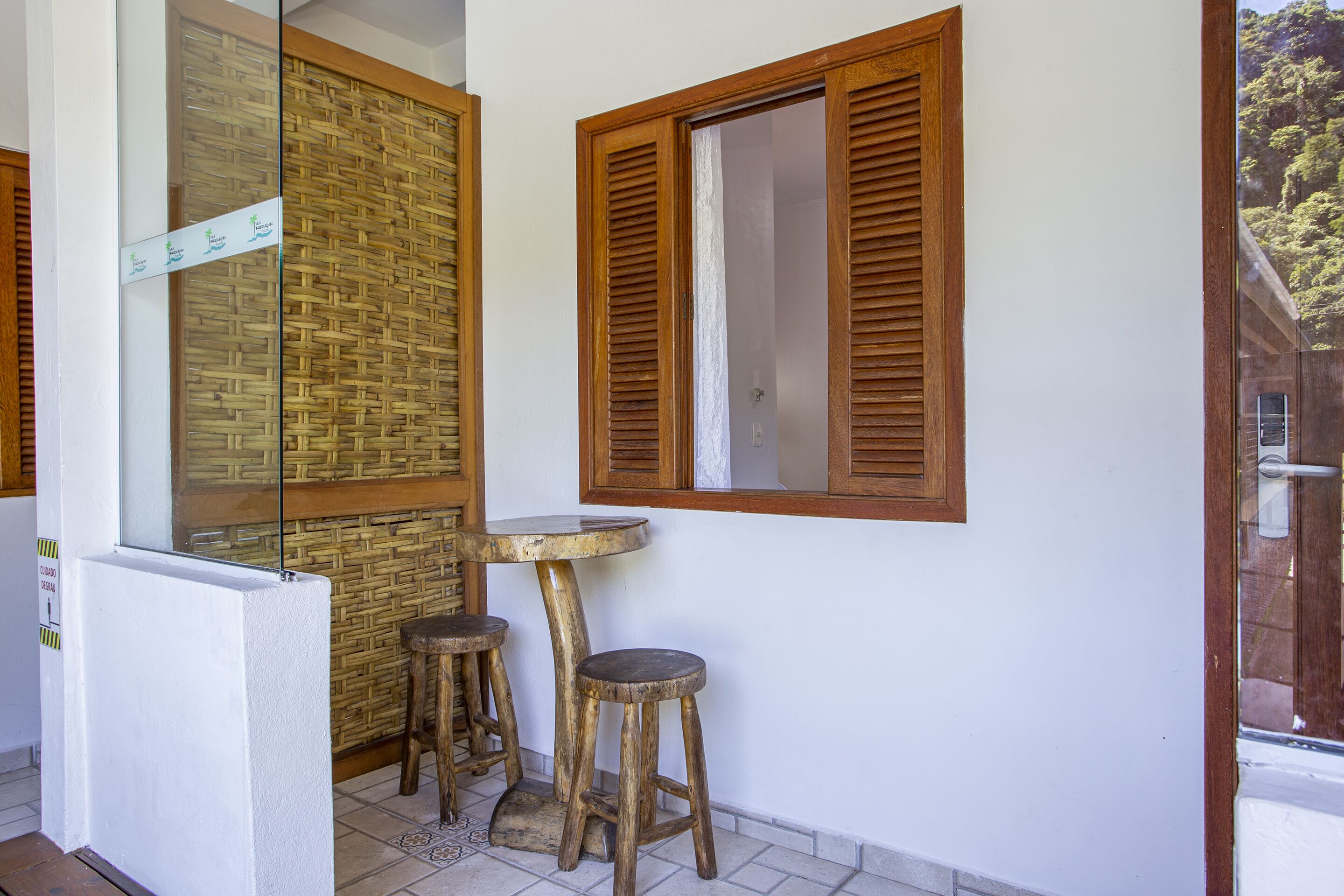 Un petit balcon avec une table en bois et deux tabourets, une cloison en verre et une fenêtre avec des volets en bois.