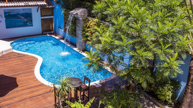 A swimming pool with a wooden deck, surrounded by lush greenery and a small waterfall feature. - Pousada Vila Barequeçaba (São Sebastião)